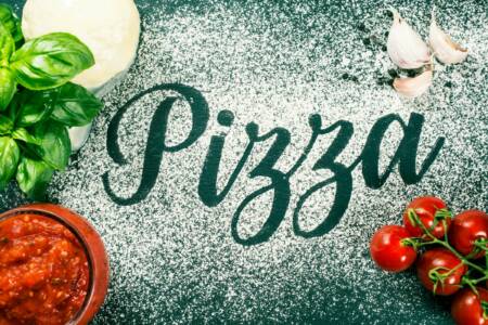 Auf dem Tisch liegt Mehl, in das der Schriftzug ‚Pizza‘ geschrieben wurde – daneben leuchten frische, rote Tomaten.